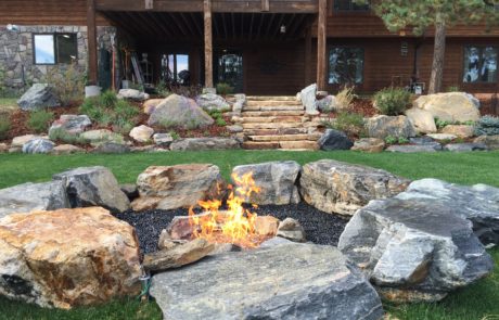 Lit fireplace with large rocks for seating, looking back at the house with other stone and plant landscaping