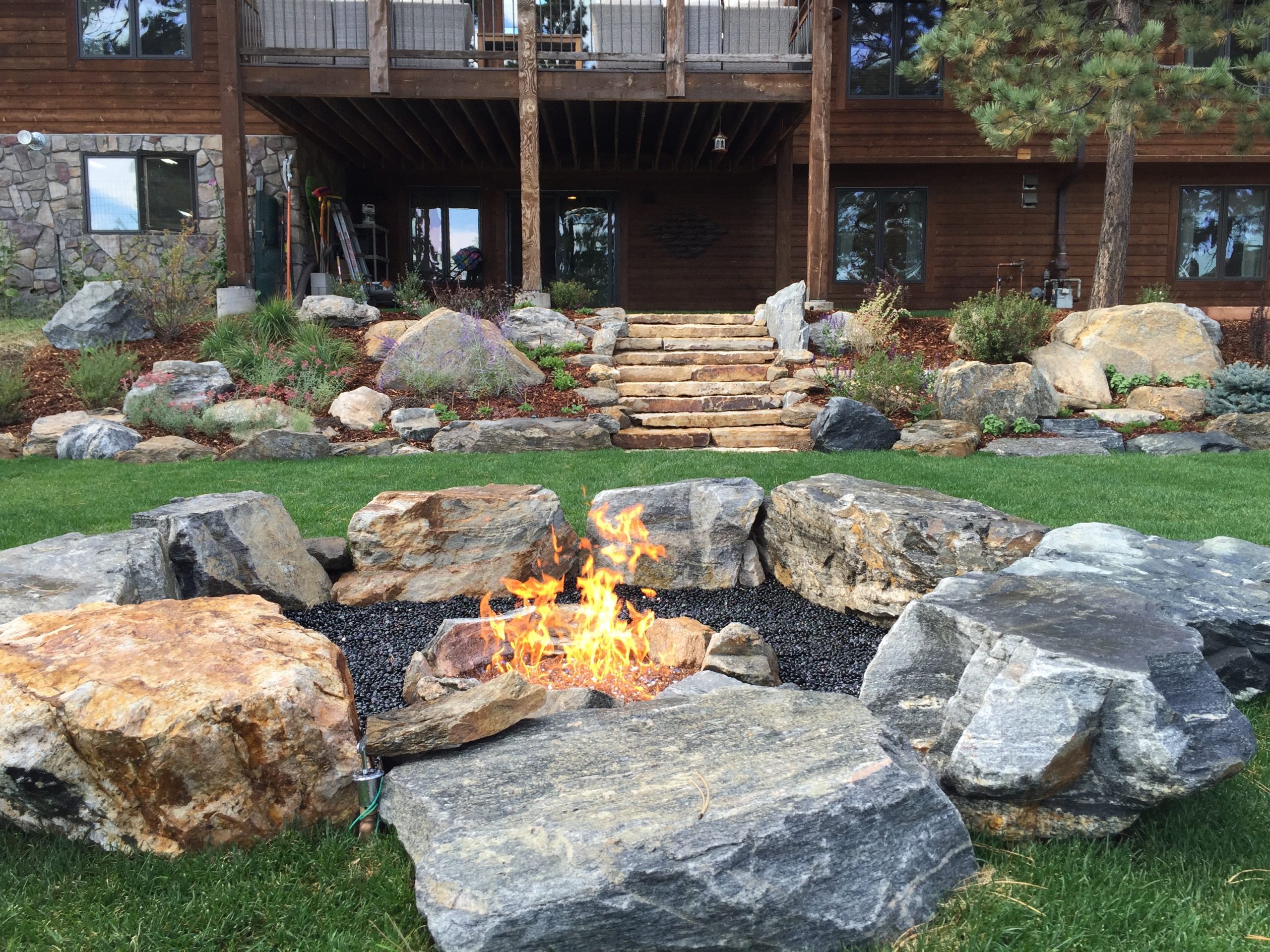 Lit fireplace with large rocks for seating, looking back at the house with other stone and plant landscaping