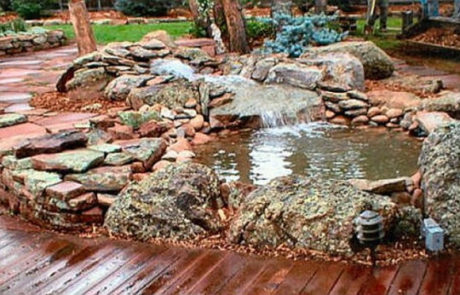 Wooden deck next to small pond and waterfall, lined with rocks of various size